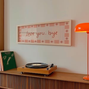 Puede incluir: Una obra de arte enmarcada con el texto "Love you. bye" en rosa, rodeada por un borde a cuadros rosa y blanco y estrellas. Un tocadiscos de madera y discos de vinilo est&aacute;n sobre un mueble de madera.