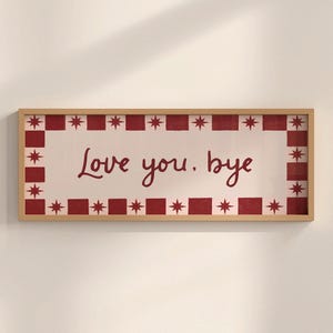 Puede incluir: Un letrero rectangular con marco de madera con las palabras "Love you. bye" en escritura burdeos. El letrero tiene un fondo blanco con un borde burdeos de cuadrados y estrellas alternos.