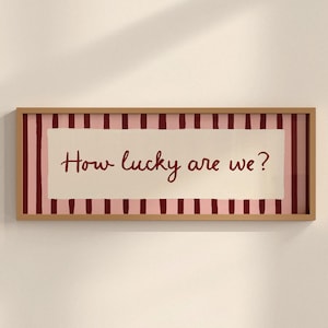 May include: A rectangular framed wall art piece featuring a light pink background with vertical dark red stripes. Centered is a cream-coloured rectangular panel with the dark red handwritten text "How lucky are we?" enclosed in a light wood frame.