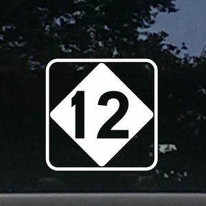May include: A white vinyl decal with a black diamond shape and the number "12" inside. The decal is on a car window.