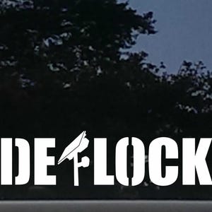 May include: A white vinyl decal applied to a dark glass surface, featuring the text DE-LOCK in a bold, stencil-style font. The letter E is replaced by a graphic illustration of a stylized lock mechanism with a small circular component.