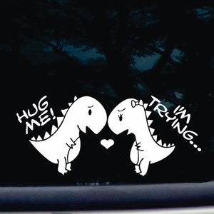 May include: White vinyl sticker featuring two cartoon dinosaurs facing each other. The dinosaur on the left says "Hug Me!" and the dinosaur on the right says "I'm Trying..."
