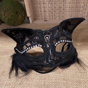 May include: Black lace cat mask with rhinestone and sequin accents. The mask features cat ears and a delicate design. A black elastic band secures the mask. The mask is suitable for costume parties or masquerades.