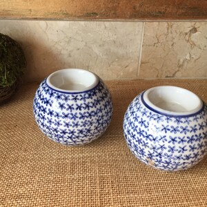 May include: Two white ceramic candle holders with a blue floral pattern. The holders are round and have a small opening at the top for a candle.