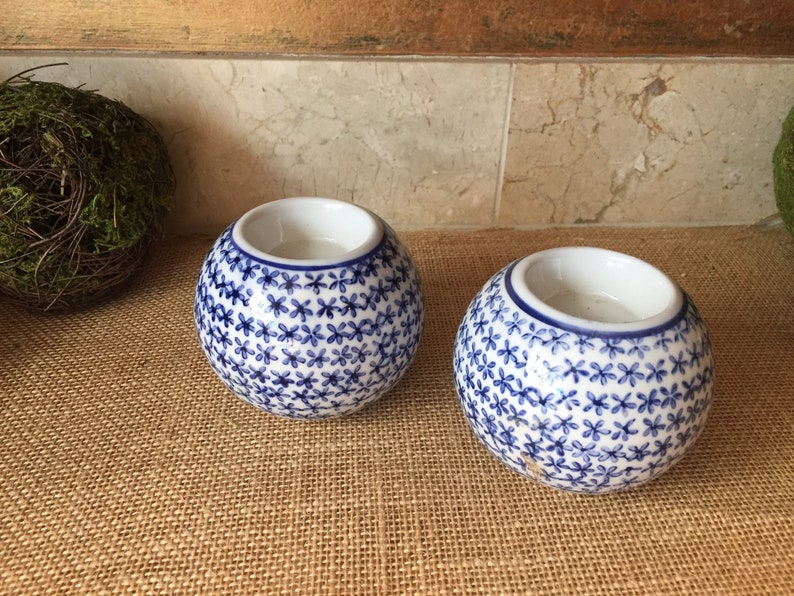 May include: Two white ceramic candle holders with a blue floral pattern. The holders are round and have a small opening at the top for a candle.