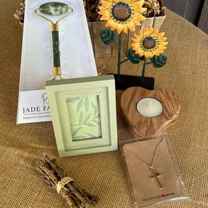 May include: A gift basket with a green jade face roller, a small picture frame with a green leaf design, a wooden heart-shaped candle holder, a gold chain necklace with a cross pendant, and a bundle of brown sticks tied with a gold ribbon.