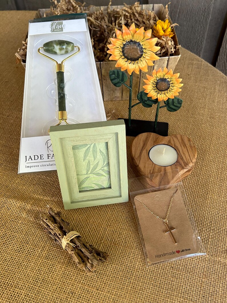 May include: A gift basket with a green jade face roller, a small picture frame with a green leaf design, a wooden heart-shaped candle holder, a gold chain necklace with a cross pendant, and a bundle of brown sticks tied with a gold ribbon.