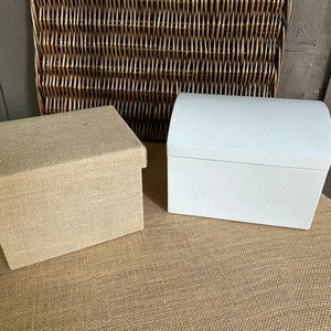 May include: Two decorative boxes, one made of natural burlap and the other white with a rounded lid. Both boxes are sitting on a brown burlap surface.