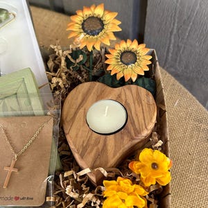 May include: A gift basket filled with brown paper shred, a wooden heart-shaped candle holder with a white candle, a gold necklace with a cross pendant, and artificial sunflowers. The basket is lined with brown burlap and has the text "made with love" on the bottom.