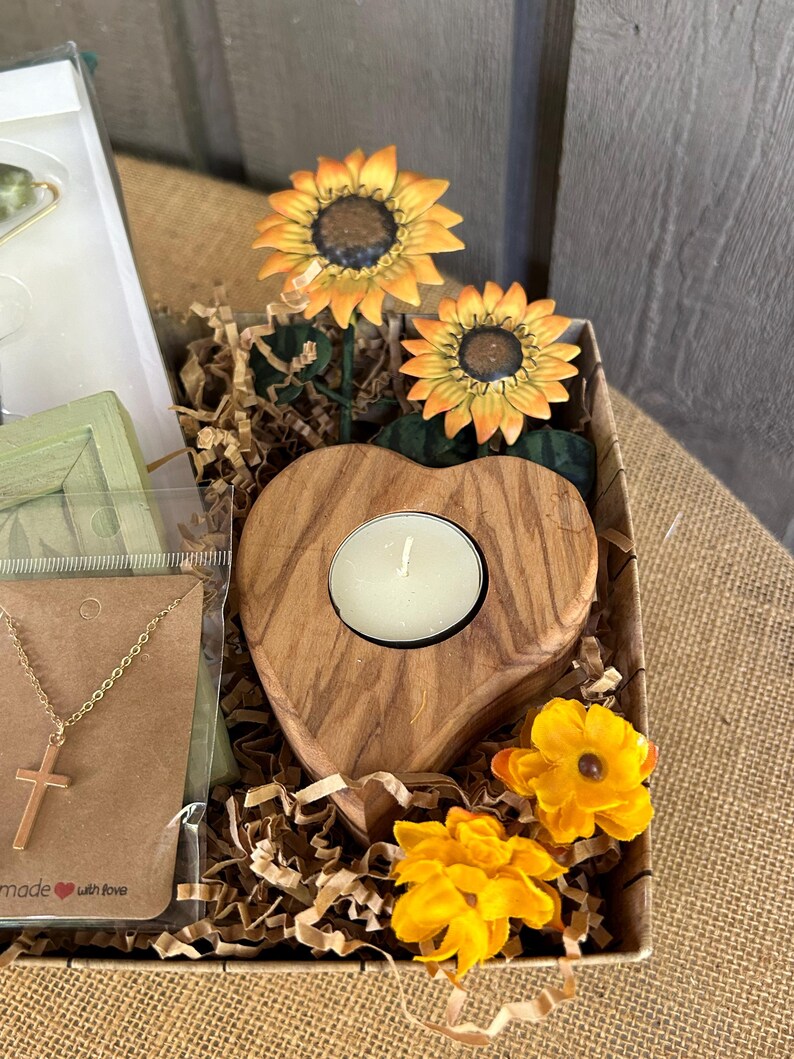May include: A gift basket filled with brown paper shred, a wooden heart-shaped candle holder with a white candle, a gold necklace with a cross pendant, and artificial sunflowers. The basket is lined with brown burlap and has the text "made with love" on the bottom.