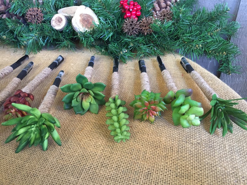 May include: A set of ten black pens decorated with faux succulents and twine. The pens are arranged on a burlap surface.