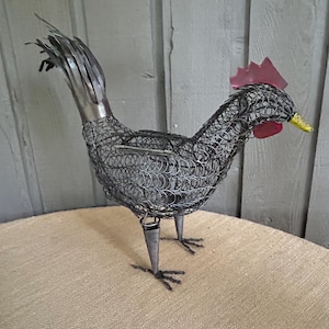 May include: A metal rooster sculpture with a wire mesh body, a red comb, and a yellow beak. The rooster stands on metal legs with wire feet. The sculpture is placed on a tan surface with a gray wooden background.