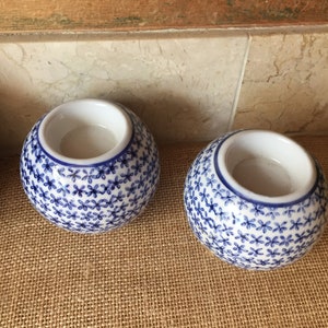 May include: Two white ceramic candle holders with a blue floral pattern. The holders are round and have a small opening at the top for a candle.