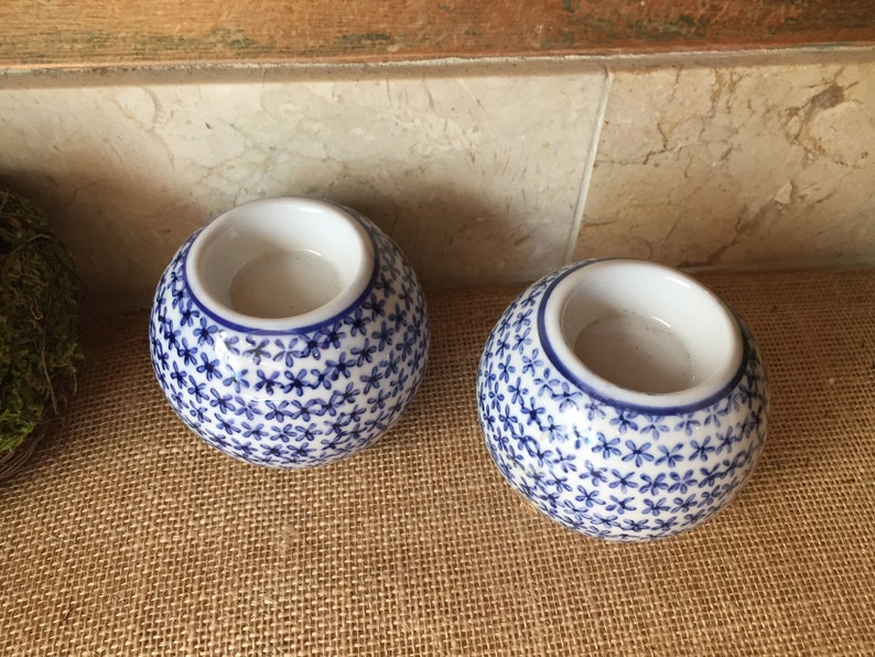 May include: Two white ceramic candle holders with a blue floral pattern. The holders are round and have a small opening at the top for a candle.