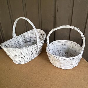 May include: Two white wicker baskets with handles. One is oval-shaped, and the other is round. Both baskets are empty and sit on a brown surface. The baskets are made of woven white wicker.