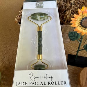 May include: A green jade facial roller with a gold handle. The roller is packaged in a white box with the text "Glam Gadgets" and "Rejuvenating Jade Facial Roller" printed on it. The text "Improve circulation for a refreshed, healthy glow" is also printed on the box.