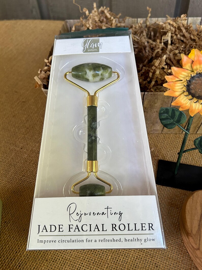 May include: A green jade facial roller with a gold handle. The roller is packaged in a white box with the text "Glam Gadgets" and "Rejuvenating Jade Facial Roller" printed on it. The text "Improve circulation for a refreshed, healthy glow" is also printed on the box.
