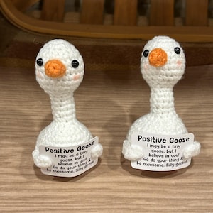 May include: Two handmade white crochet goose figurines with orange beaks and black eyes. Each goose holds a sign that reads "Positive Goose" and a motivational message. The background is a light brown surface.