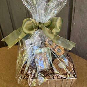 May include: A brown wicker basket filled with a variety of gifts, including a small wooden box with a cross, a candle, and a sunflower. The basket is wrapped in clear cellophane and tied with a green ribbon.