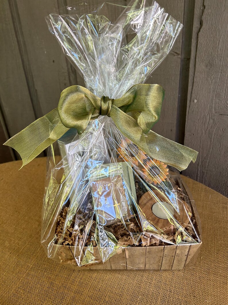 May include: A brown wicker basket filled with a variety of gifts, including a small wooden box with a cross, a candle, and a sunflower. The basket is wrapped in clear cellophane and tied with a green ribbon.