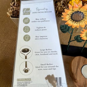 May include: A white and green package for a jade facial roller. The package features a sunflower design and text that reads "Rejuvenating Jade Facial Roller". The text also lists the benefits of using a jade roller, including reducing under-eye puffiness, tightening and reducing pores, and improving skin elasticity. The package also shows the two different sizes of rollers, a large roller and a small roller.