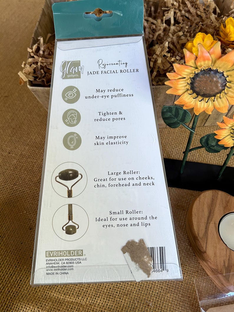 May include: A white and green package for a jade facial roller. The package features a sunflower design and text that reads "Rejuvenating Jade Facial Roller". The text also lists the benefits of using a jade roller, including reducing under-eye puffiness, tightening and reducing pores, and improving skin elasticity. The package also shows the two different sizes of rollers, a large roller and a small roller.