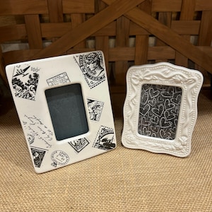 May include: A rectangular white picture frame featuring black postage stamp graphics and a smaller, ornate white picture frame with a raised floral border and a black insert printed with a white heart pattern, both standing upright.