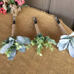 May include: Three pens decorated with faux greenery and twine. The pens are wrapped in twine and have a small bouquet of green and gray leaves attached to the top.