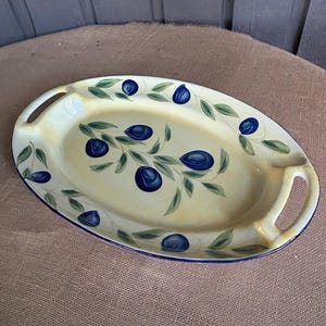 May include: An oval ceramic serving platter with a cream-colored base and a hand-painted pattern of dark blue plums and green leaves. The platter features two integrated handles on the sides and a thin blue rim around the outer edge.