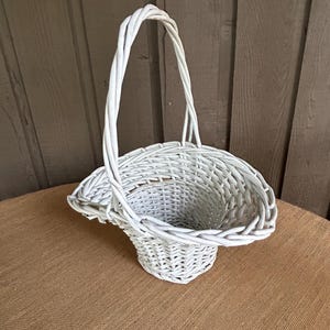 May include: A white wicker basket with a curved handle. The basket has a flared shape and is made of woven white wicker. The basket is sitting on a brown surface.