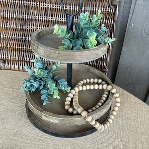 May include: Two-tier wooden serving tray with a black metal stand. The top tier has a small green plant and the bottom tier has a strand of wooden beads.