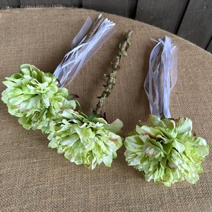 May include: Three green silk flower wands with white ribbon streamers. The wands are tied with a white ribbon and have a wooden stick base.