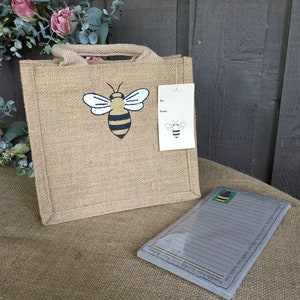 May include: A brown burlap tote bag with a bee design. The bag has a gift tag attached with the words "To" and "From". The bag is sitting on a burlap surface with a gray notepad with a bee design.