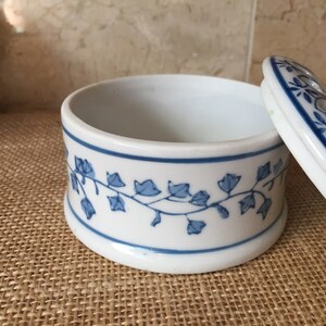 May include: A white ceramic trinket box with a blue and white floral design. The lid has a small hole in the center and is open, revealing the empty interior of the box.