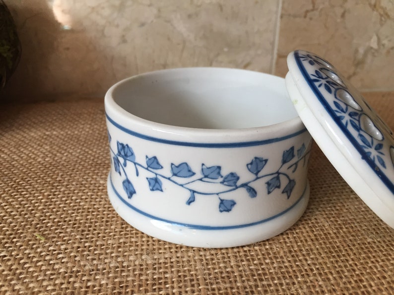 May include: A white ceramic trinket box with a blue and white floral design. The lid has a small hole in the center and is open, revealing the empty interior of the box.