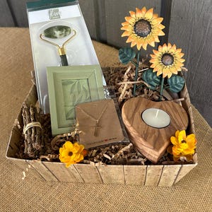 May include: A gift basket filled with items including a green jade roller, a small picture frame, a gold necklace with a cross pendant, a wooden heart-shaped candle holder, and artificial sunflowers.