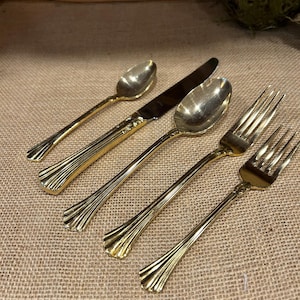 May include: A set of gold-colored flatware, including a spoon, knife, spoon, fork, and fork. The handles have a decorative, fluted design. The utensils are arranged on a textured, tan surface.