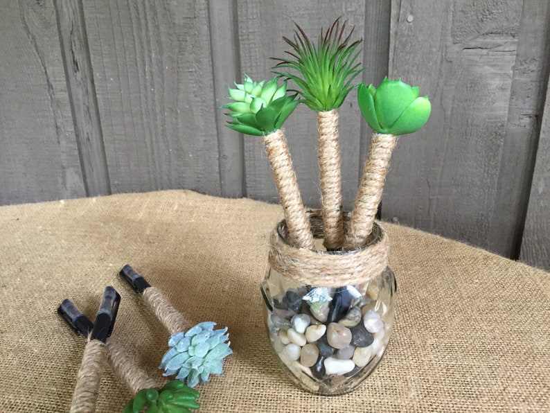 May include: Three faux succulent plants in a glass jar with a burlap rope wrapped around the top. The jar is filled with small gray and white stones. The plants are in a burlap-covered vase.