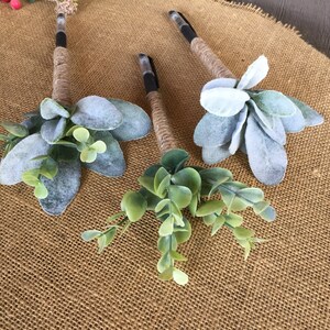 May include: Three pens wrapped in twine and decorated with faux greenery. The pens are decorated with sprigs of eucalyptus and lambs ear.