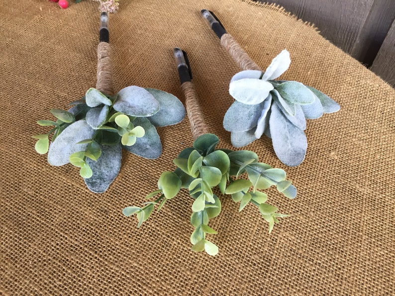 May include: Three pens wrapped in twine and decorated with faux greenery. The pens are decorated with sprigs of eucalyptus and lambs ear.