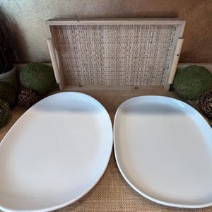 May include: Two white oval ceramic serving platters on a burlap surface. The platters are slightly raised in the center and have a smooth, glossy finish.