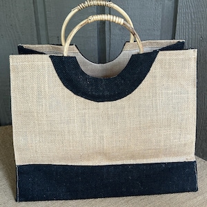 May include: A rectangular jute tote bag with a natural tan body and black accents. The bag features a black band at the bottom and a curved black trim at the top. It has two bamboo handles.