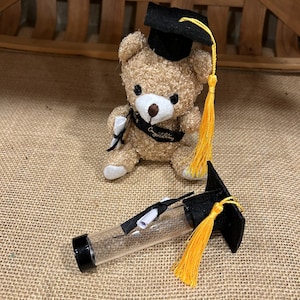 May include: A small, tan teddy bear wearing a black graduation cap with a yellow tassel and a ribbon that says "Congratulations." A clear tube with a black cap and a miniature scroll is next to the bear. Graduation themed.