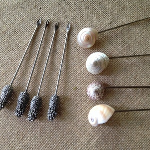 May include: A set of eight metal cocktail picks. Four picks feature silver-toned, textured, pinecone-shaped handles with two-pronged tips. The other four picks feature natural, spiral-shaped seashells attached to the ends of thin, straight metal shafts.