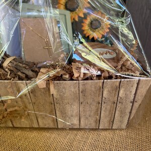 May include: A wooden gift basket filled with brown tissue paper, a gold necklace with a cross pendant, a small framed picture, and two artificial sunflowers.