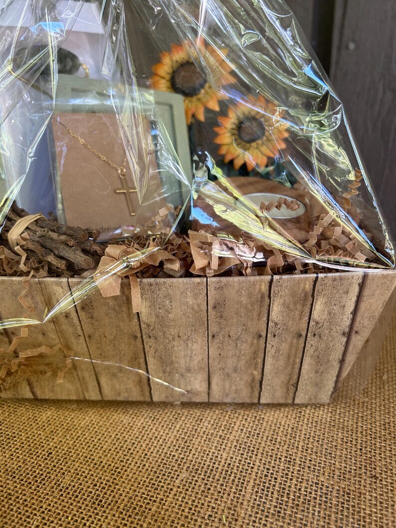 May include: A wooden gift basket filled with brown tissue paper, a gold necklace with a cross pendant, a small framed picture, and two artificial sunflowers.