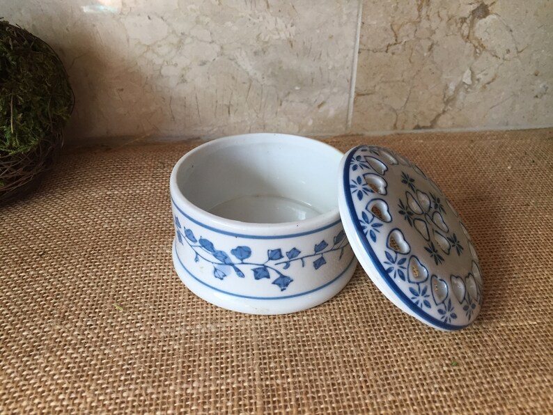 May include: A white ceramic trinket box with a blue and white floral design. The lid has a cutout design and is open.