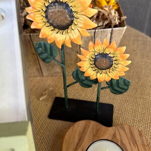 May include: Two metal sunflowers with green leaves on a black stand. A wooden heart-shaped candle holder with a white tea light candle.