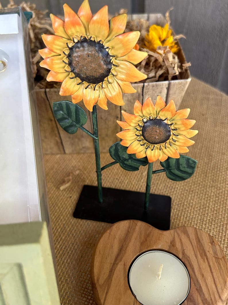 May include: Two metal sunflowers with green leaves on a black stand. A wooden heart-shaped candle holder with a white tea light candle.