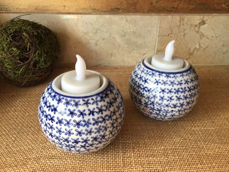 May include: Two white ceramic candle holders with blue floral patterns. Each holder has a white LED tea light inside.